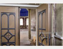 Holzflügeltüren mit geometrischen und kreuzförmigen Verzierungen führen in einen leicht verfallenen, historisch anmutenden Innenraum mit bogenförmigen Fenstern, blau gestrichener Kuppeldecke und einfachen Möbeln.