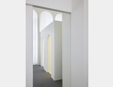 Minimalistischer Innenraum mit weißen Wänden, hohen gewölbten Durchgängen und einem grauen Bodenbelag. Im Hintergrund sind bodentiefe, lichtdurchflutete Vorhänge sichtbar.