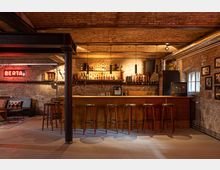 Eine rustikale Bar mit gemauerter Backsteinwand, beleuchtet durch warme Pendelleuchten. Die Bar verfügt über mehrere Holzhocker, dekorative Cowboy-Stiefelregale und eine Flasche-Auslage; im Hintergrund ein rotes Neonlicht mit der Aufschrift "BERTA".