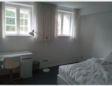 Helles Schlafzimmer mit zwei kleinen Fenstern mit Sprossen und weißen Vorhängen. Im Raum befinden sich ein Schreibtisch mit weißem Stuhl und Stehlampe sowie ein ungemachtes Bett mit weißer Bettwäsche.