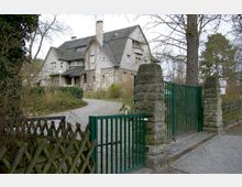 Steinernes Wohnhaus mit Fachwerkelementen und dunklem Schrägdach, umgeben von Bäumen und Sträuchern. Im Vordergrund ein gepflasterter Weg, ein grünes Metalltor mit steinernen Pfeilern und ein Holzzaun.