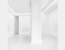 Minimalistischer, weißer Innenraum mit glattem Boden und kastenförmiger Raumstruktur; im Vordergrund eine säulenartige Struktur mit feinen vertikalen Rillen, die die Decke stützt. Die Decke hat eine eingefasste Beleuchtung, die den Raum gleichmäßig erhellt.