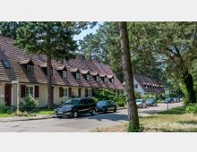 Reihenhäuser mit rotem Ziegeldach und kleinen Gaubenfenstern, umgeben von hohen Bäumen an einer ruhigen Wohnstraße mit geparkten Autos.