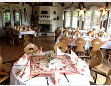 Gemütlicher Speisesaal im Landhausstil mit Holzstühlen, weiß gedeckten Tischen, roten und weißen Servietten sowie einem Kachelofen im Hintergrund. Große Fenster und rustikale Dekorationen wie Ski und landwirtschaftliche Werkzeuge verleihen dem Raum eine traditionelle Atmosphäre.