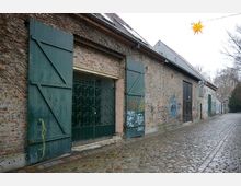 Eine gepflasterte Gasse mit einer langen Backsteinfassade, an der mehrere grüne Holztüren und Gitterfenster angebracht sind. Die Wand ist teils mit Graffiti verziert, und im Hintergrund hängt ein gelber Stern als Dekoration.