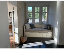 Gemütlicher Wohnbereich mit einer weißen Bank mit Kissen und einem Fenster im Erkerstil, das viel Tageslicht hereinlässt. Rechts neben der Bank steht ein Korb mit Decken, während links ein Durchgang zur Küche mit weißen Fliesen und Holzelementen führt.