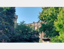 Backstein-Gebäude mit großen, rechteckigen Fenstern, umgeben von dichten grünen Bäumen, bei klarem Himmel fotografiert. Im unteren Bereich ist ein kleiner überdachter Eingang mit Graffiti sichtbar.