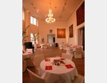 Eleganter Veranstaltungsraum mit hohen Decken, großem Kronleuchter und großen Fenstern, die viel Tageslicht hereinlassen. Rund eingedeckte Tische mit weißem Tischtuch und Dekoration, umgeben von Kunstwerken an den cremefarbenen Wänden.