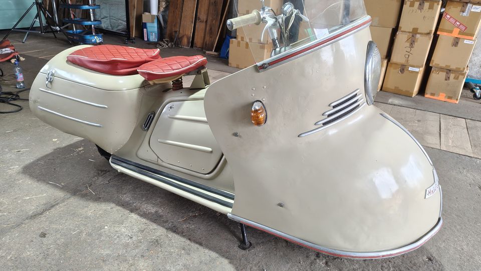 Innenraum einer Werkstatt oder Lagerhalle mit Betonboden, gestapelten Kartons und Holzplatten im Hintergrund. Im Vordergrund steht ein cremefarbenes Vintage-Kleinfahrzeug mit großer Frontverkleidung, Windschutzscheibe und zwei roten Sitzpolstern.