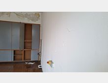 Leerer Innenraum mit abblätternder Deckenfarbe und stark abgenutztem Holzboden; an der linken Wand steht ein eingebauter Schrank mit teils offenen Türen und Regalböden. An der rechten Wand befinden sich Risse im Putz sowie ein Not-Aus-Schalter.