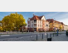 Straßenansicht mit einem historischen Gebäude aus rotem Backstein, das über helle Fensterrahmen und ein rotes Ziegeldach verfügt. Im Vordergrund sind eine gepflasterte Straße und ein Platz zu sehen, während im Hintergrund Wohnhäuser und ein großer Baum mit herbstlich gelbem Laub stehen.