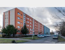 Mehrstöckige Wohngebäude in Plattenbauweise, teilweise in orange und blau gestrichen, entlang einer ruhigen Straße mit parkenden Autos. Im Vordergrund sind Gehwege, Grünflächen und vereinzelt Bäume sichtbar, im Hintergrund ist eine Kirchturmspitze erkennbar.