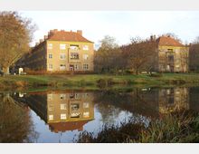 Wohngebäude mit roten Ziegeldächern und beige gestrichenen Fassaden, umgeben von Bäumen in einer herbstlichen Parklandschaft, spiegeln sich in einem ruhigen Teich im Vordergrund. Die Umgebung umfasst Rasenflächen, Büsche und Bänke.