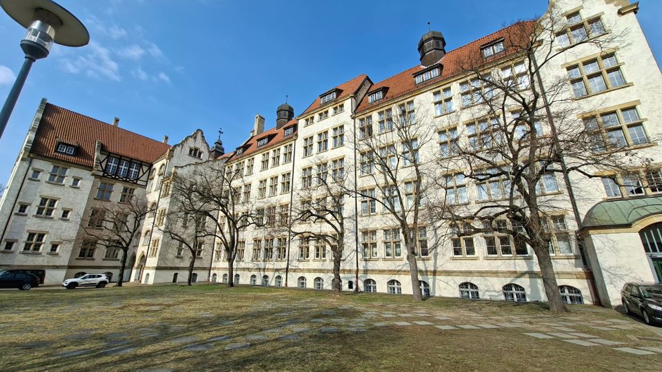 Großer Innenhof vor einem mehrstöckigen historischen Gebäude mit heller Fassade, vielen rechteckigen Fenstern und roten Ziegeldächern mit kleinen Dachaufbauten. Im Vordergrund stehen kahle Bäume und eine gepflasterte Rasenfläche, links ist ein Flügel mit Fachwerkdetails zu sehen.