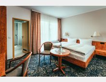 Hotelzimmer mit einem Doppelbett, hellem Holzmobiliar, einem kleinen runden Tisch und zwei Stühlen auf einem gemusterten Teppichboden. Im Hintergrund befindet sich ein Fenster mit Vorhängen sowie eine offene Tür zum Badezimmer mit Dusche.