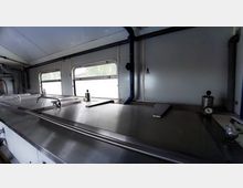 Innenraum eines industriellen Arbeitsbereichs mit Edelstahl-Arbeitsflächen und integrierten Deckeln. Im Hintergrund befinden sich mehrere Fenster mit Sicht nach draußen sowie Rohrleitungen und eine Wand mit Beschriftung.