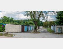 Reihe kleiner Garagen mit verschiedenfarbigen Türen, flankiert von Bäumen und Büschen, auf einem gepflasterten Gelände mit einem Straßenschild und einem Baum im Vordergrund. Im Hintergrund ist ein Funkmast sichtbar.