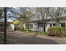 Innenhof mit gepflasterter Fläche und mehreren kahlen Bäumen vor einem eingeschossigen Gebäude mit flachem Dach, Fenstern und einer farbig gerahmten Eingangstür. Am Rand stehen Sträucher und kleine Grünflächen, links ist ein überdachter Bereich mit Wand und Sitzgelegenheit zu sehen.