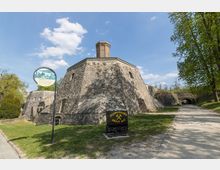 Historisches Steingebäude in einem Museumsgelände, umgeben von Rasenflächen und Bäumen, mit einem schmalen Fußweg neben dem Gebäude. Ein Schild mit der Aufschrift "Museumspark Glück auf!" sowie ein kleiner Förderwagen befinden sich im Vordergrund.