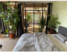 Schlafzimmer mit Doppelbett und grauer Bettwäsche, flankiert von Zimmerpflanzen und einem Nachttisch. Im Hintergrund befindet sich eine Glastrennwand mit schwarzen Rahmen, die den Blick in einen hellen Wohnraum mit Holzboden und weiteren Pflanzen eröffnet.