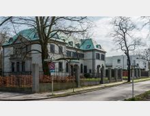 Eine Villa im historischen Stil mit hellen Fassaden und grünem Mansarddach, umgeben von einem schmiedeeisernen Zaun und einer gepflegten Gartenanlage. Im Vordergrund befindet sich ein Gehweg mit Straßenschildern und kahlen Bäumen.