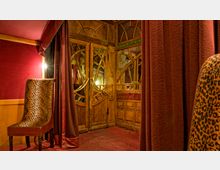 Innenansicht eines Raumes mit opulentem Design, geprägt von roten Vorhängen, Holztüren mit verzierten Glaseinsätzen und einem gepolsterten Stuhl mit Leopardenmuster. Der Raum hat warme Beleuchtung und eine rote Teppichbodenverkleidung.