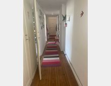 Ein schmaler Flur in einer Wohnung mit Holzdielenboden und weißen Wänden. An der Wand hängen Bilderrahmen, dekorative Elemente und Aufbewahrungsregale, während bunte, gestreifte Teppiche den Boden bedecken; am Ende des Flurs ist eine helle Tür zu sehen.