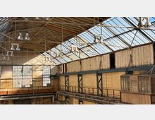 Innenansicht einer ehemaligen Industriehalle mit hohem Glasdach auf Stahlträgern, hängenden Hallenleuchten und großen Sprossenfenstern. Ziegelwände, umlaufende Stahlkonstruktionen und sichtbare Rohrleitungen prägen den Raum.