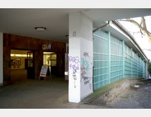 Eingangsbereich einer U-Bahn-Station mit einem Wegweiser für den U-Bahn-Zugang. Links ein Zugang mit Glastüren, rechts ein langes, türkisfarbenes Gitter und eine Steinmauer mit Graffiti.