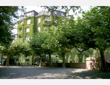 Mehrstöckiges Gebäude mit komplett von grünem Efeu bewachsener Fassade, umgeben von schattenspendenden Bäumen auf einem Platz mit hellem Kiesboden und Steinmauern im Vordergrund.