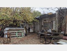 Hinterhof mit chaotisch gelagerten Gegenständen, darunter Holzpaletten, Metallrahmen, Tische und Stühle. Im Hintergrund stehen ein altes, verwittertes Gebäude mit grauen Ziegeln und eine Überdachung, während Bäume und Laub die Umgebung prägen.