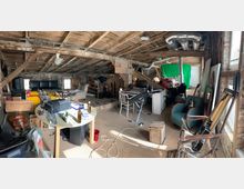 Ein unaufgeräumter Dachboden mit sichtbaren Holzbalken und unverputzten Ziegelwänden. Der Raum enthält Möbel wie Sessel, Tische und Stühle, Musikinstrumente wie Gitarren, sowie einen Computerarbeitsplatz und verschiedenes Studioequipment wie ein Mikrofon und eine grüne Leinwand.