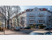 Mehrstöckiges Wohngebäude mit heller Fassade und roten Ziegeldächern, an einer Straßenecke mit Gehweg und parkenden Autos. Im Erdgeschoss befindet sich ein Biergarten mit einem grünen Vordach, umgeben von kahlen Bäumen in einer urbanen Umgebung.