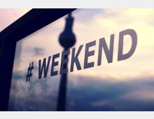 Eine Glasfläche mit dem Schriftzug "#WEEKEND", im Hintergrund verschwommen die Silhouette eines hohen Turms vor einem bewölkten Himmel.
