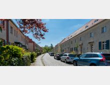 Straßenszene in einem Wohngebiet mit Reihenhäusern auf beiden Seiten, teils mit pastellfarbenen Fassaden und Ziegeldächern. Eine mit Autos gesäumte Kopfsteinpflasterstraße führt durch die Szenerie, flankiert von Grünflächen und Bäumen.