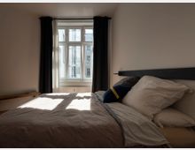 Schlicht eingerichtetes Schlafzimmer mit einem großen Bett, darüber ein schlichtes schwarzes Kopfteil. Im Hintergrund befindet sich ein Fenster mit weißen Vorhängen und dunklen Übergardinen, durch das Tageslicht einfällt.