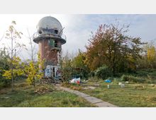 Ein verlassener, runder Backsteinturm mit einer großen, grauen Kuppel, teilweise mit Graffiti bedeckt, steht in einer überwucherten, unordentlichen Grünfläche. Im Vordergrund sind hohe Gräser, Bäume mit Herbstlaub und verstreute Gegenstände sichtbar.