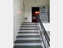 Treppenaufgang in einem öffentlichen Gebäude mit einem Metallgeländer auf der rechten Seite. Am oberen Ende der Treppe führt eine offene Tür zu einem Raum mit dunklerer Beleuchtung und Plakaten an den Wänden.