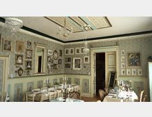 Ein nostalgisch gestalteter Speiseraum mit dekorativen, detailreichen Wänden, die mit zahlreichen gerahmten Bildern und Spiegeln verziert sind. Der Raum hat hohe Decken mit Kronleuchtern, gedeckte Tische mit Kerzenleuchtern und ein geöffneter Durchgang zu einem weiteren Raum.