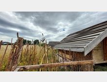 Rustikaler Außenbereich mit einem kleinen Gebäude mit Holzdach und Lehmwänden. Im Vordergrund befindet sich ein einfacher Zaun aus ungeschält bearbeiteten Ästen, umgeben von hohem Gras und Feldpflanzen, unter einem bewölkten Himmel.
