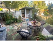 Ein kleiner Garten mit einer gemütlichen Sitzgruppe bestehend aus einem runden Tisch und Stühlen, umgeben von Pflanzen und Blumen. Im Hintergrund steht ein hellgraues Gartenhaus mit einer weißen Tür und Terrasse, während der gepflasterte Boden in bunten Steinplatten gestaltet ist.