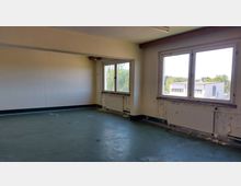 Ein leerer, renovierungsbedürftiger Raum mit abgenutzten Wänden und einem grünen Bodenbelag. Zwei große Fenster mit Blick auf draußen lassen Tageslicht herein, unter den Fenstern befinden sich Heizkörper.