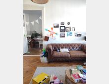 A cozy living room with a vintage brown leather sofa against a white wall adorned with framed pictures and colorful artwork. A wicker coffee table holds books and items, and an open doorway reveals a dining area with wooden furniture and indoor plants.