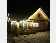 Ein Einfamilienhaus in einer winterlichen Umgebung, beleuchtet durch zahlreiche weiße Lichterketten an der Dachkante. Der Vorgarten ist leicht verschneit, und durch die Fenster ist ein beleuchteter Innenraum zu sehen.