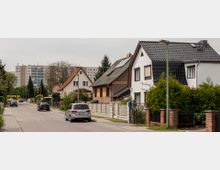 Eine Wohnstraße mit Einfamilienhäusern, teils mit roten oder dunklen Ziegeldächern, und gepflegten Vorgärten; im Hintergrund ein mehrstöckiges Wohngebäude. Autos sind am Straßenrand geparkt, und einige Gehsteiglampen säumen die Straße.