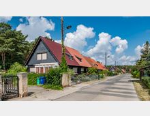 Reihen von Einfamilienhäusern mit roten und grauen Satteldächern entlang einer ruhigen Wohnstraße, umgeben von Gärten und Bäumen, bei klarem Himmel mit einigen Wolken.