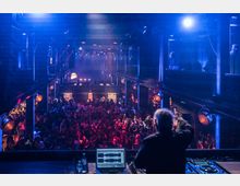 Eine Innenansicht einer dunklen Konzert- oder Clubhalle, beleuchtet mit blauen und roten Scheinwerfern. Im Vordergrund steht ein DJ an einem Mischpult, während sich eine große Menschenmenge auf einer unteren Ebene versammelt.