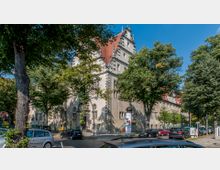 Historisches Gebäude mit hellem Putz und rotem Walmdach, verziert mit dekorativen Giebeln und Fenstern, umgeben von Bäumen und geparkten Autos auf einer gepflasterten Straße. Im Vordergrund sind Laternenmasten und ein Infokasten zu sehen.