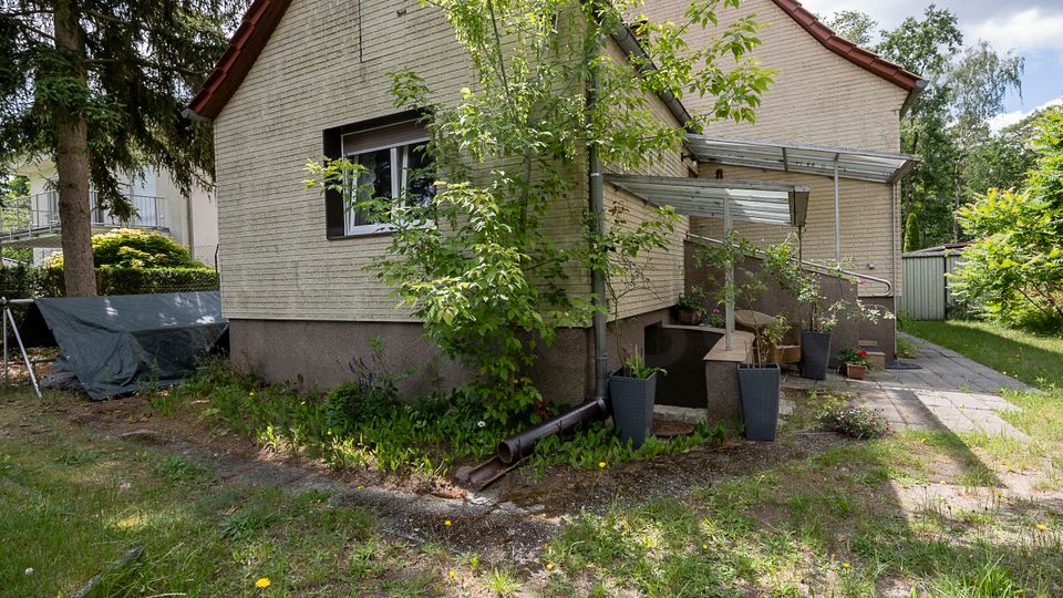 Seitenansicht eines kleinen Backsteinhauses mit Satteldach und Fenster, daneben ein überdachter Eingangsbereich mit Glasdach und Handlauf. Im Vordergrund liegen ein Garten mit Gras, Bäumen und Sträuchern sowie ein gepflasterter Weg und mehrere Pflanzkübel.