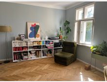 Wohnzimmer mit hellblauen Wänden, einem hölzernen Fischgrätenparkettboden und einem großen weißen Regal voller Bücher. Links steht eine Stehlampe, rechts ein grüner Sessel vor einem Fenster mit weißen Rahmen. Zimmerpflanzen und dekorative Objekte, darunter ein abstraktes Kunstbild, ergänzen die Einrichtung.